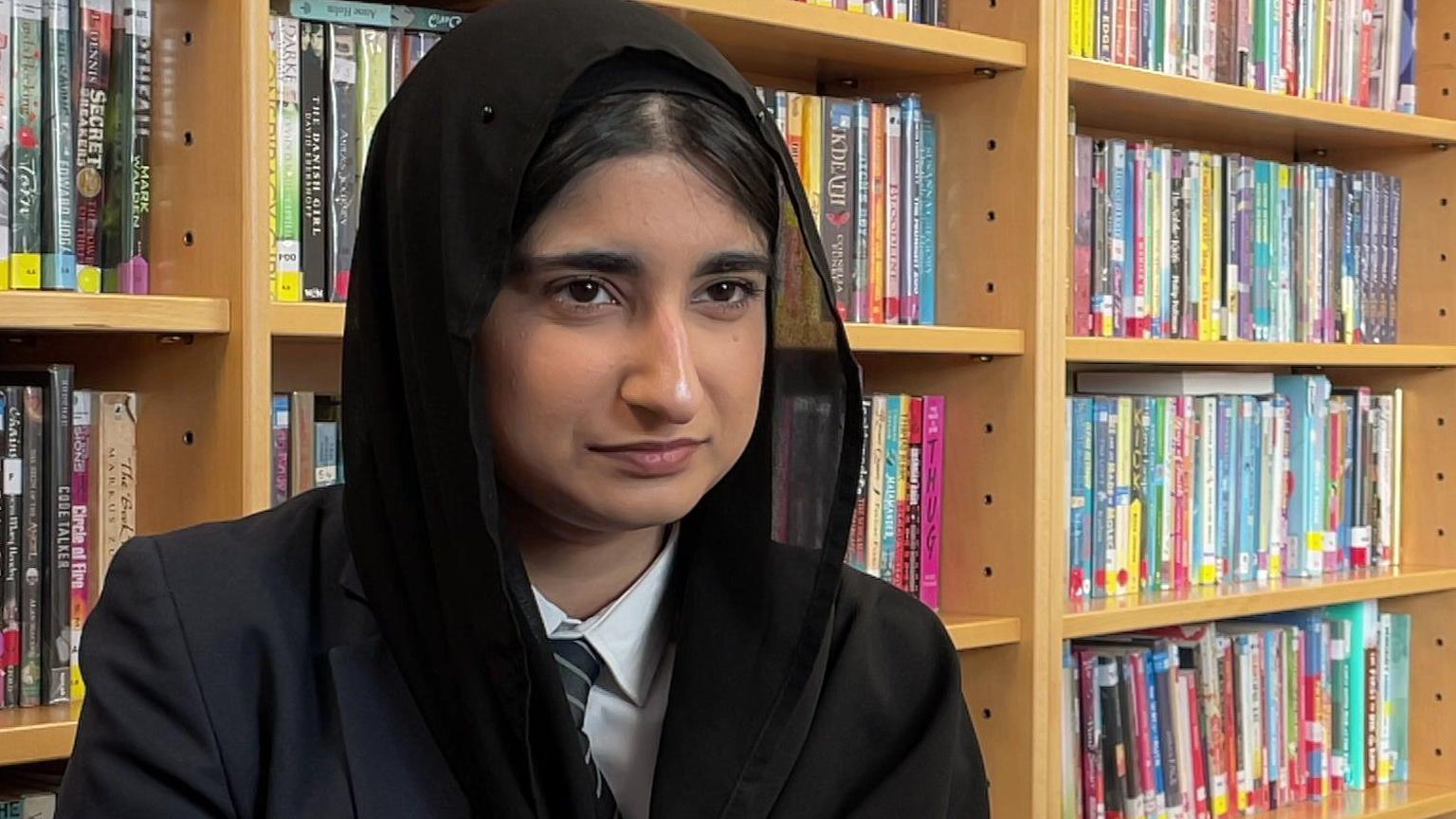 A teenage girl wearing a black headscarf sat next to a librarby shelf full of books