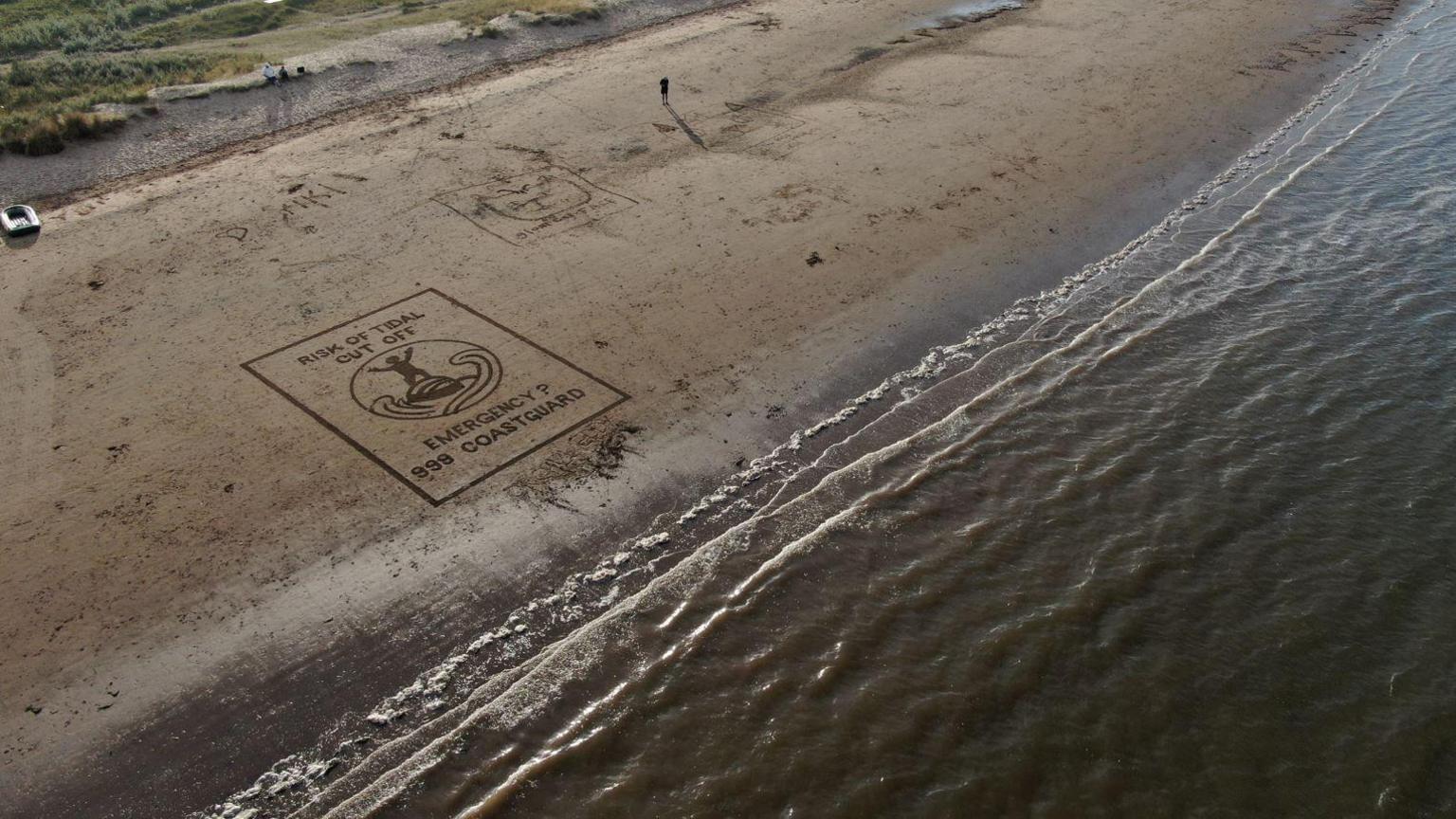 An aerial view of a stretch of beach and the sea, with the art on the sand. A rectangular box has the writing "Risk of tidal cut off. Emergency? 999 Coastguard" in it.