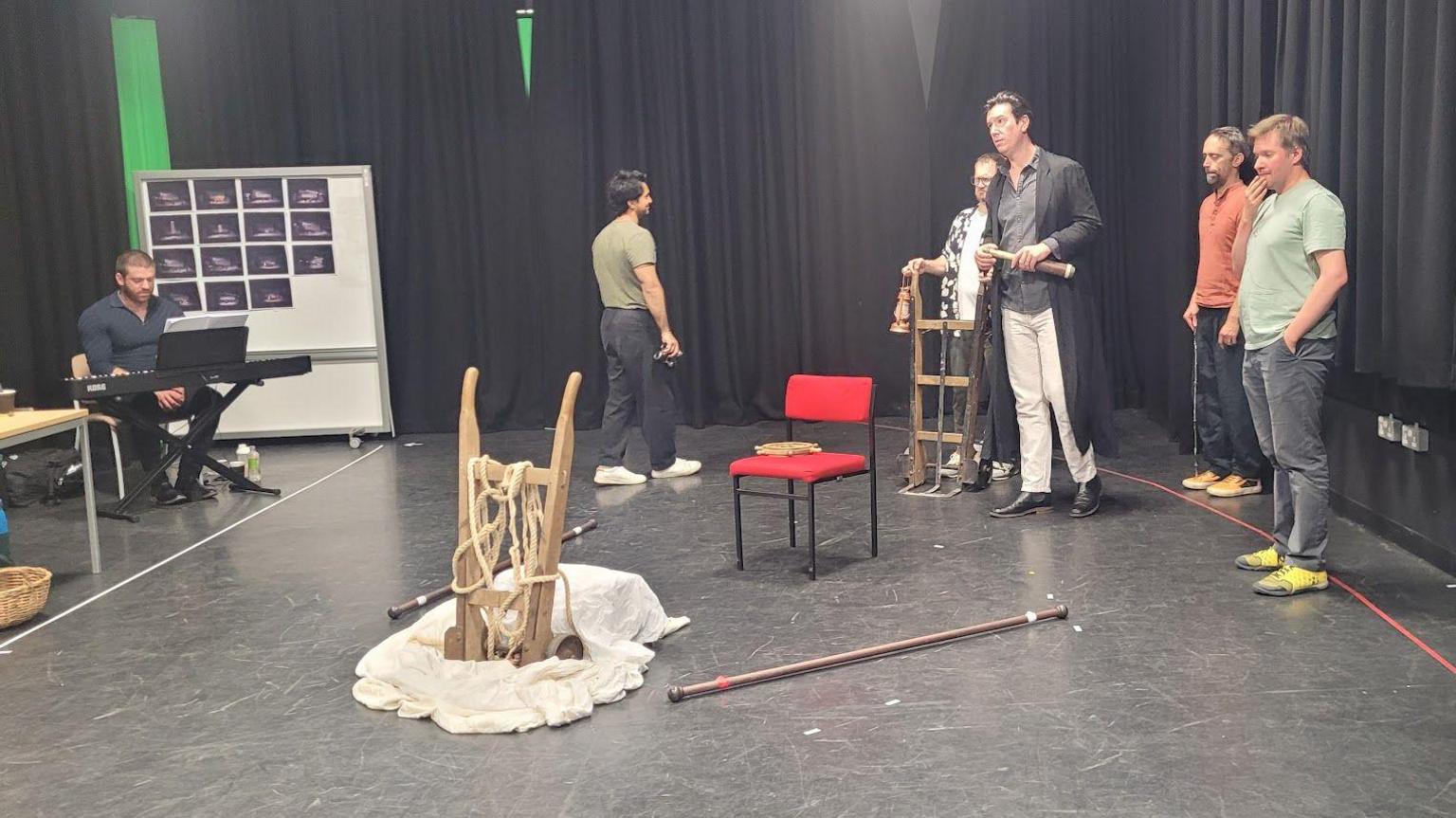 Lliam Paterson sits at a keyboard to the left of the rehearsal space while five cast members stand to the right. Various props including a red chair are in front of them 