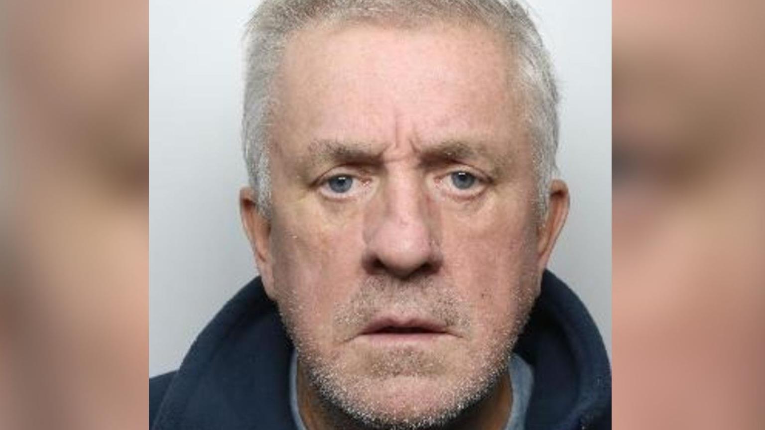 A police custody picture of a man staring straight ahead with a neutral expression. He has short, grey hair and a grey stubble and is wearing a blue jacket.