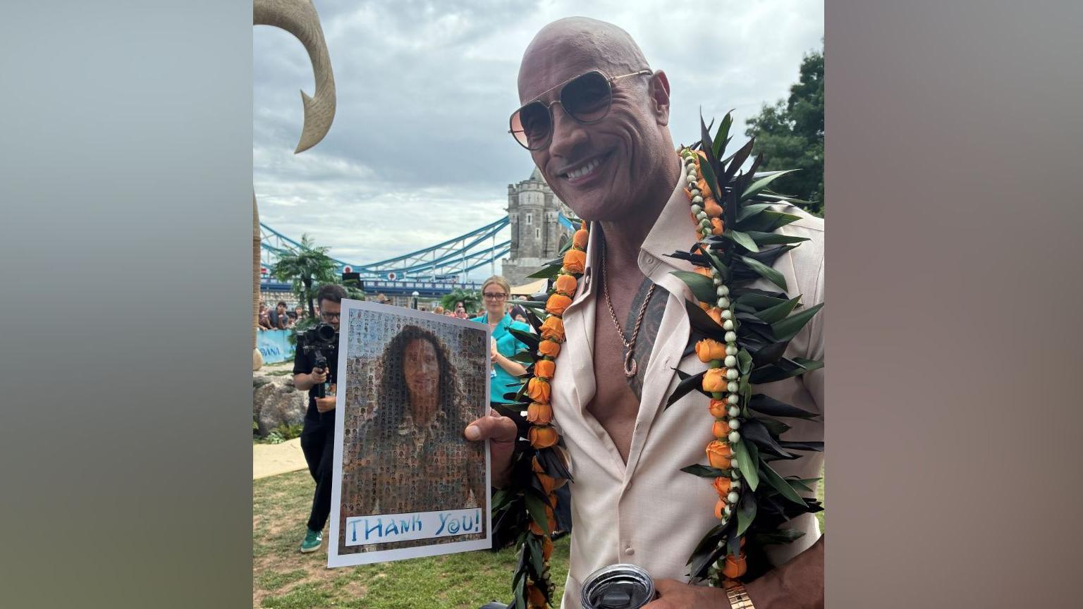 The Rock – a man wearing a cream-coloured shirt and flowers around his neck. He is also wearing sunglasses and holding up a picture of himself as the character Maui. He is smiling.