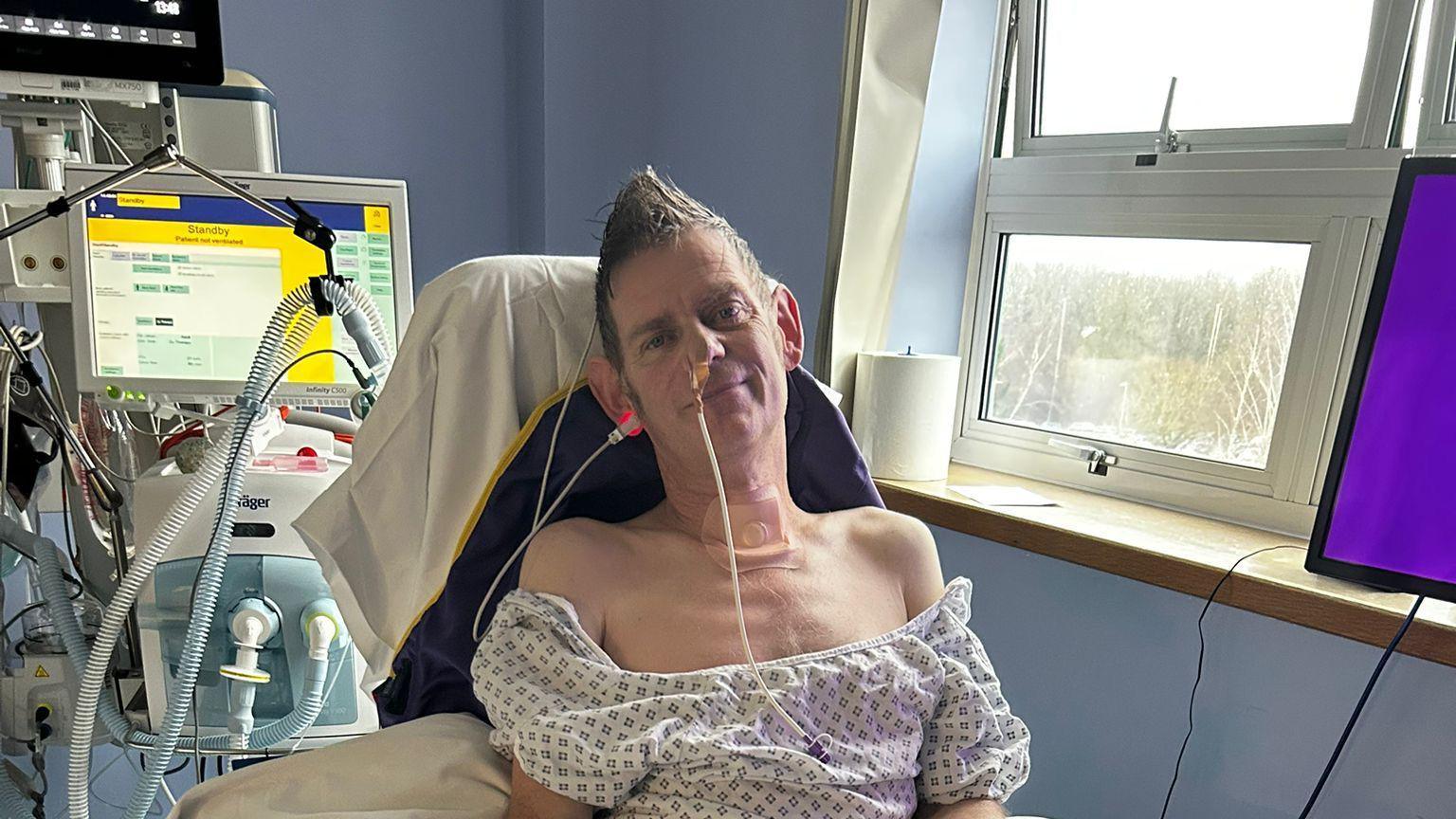 A man sitting up in a hospital bed with tubes sticking out of his nose and ears with a medical machine behind him.