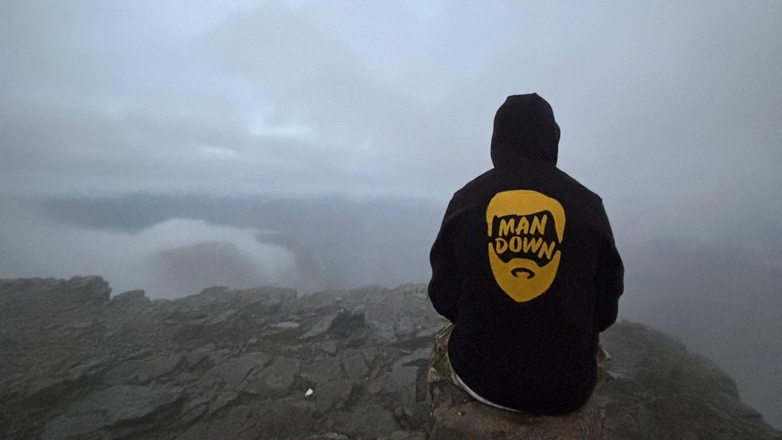 A person sitting on a rock looking into the foggy distance. They are wearing a black hoodie with the hood up. A yellow logo on the back says man down.