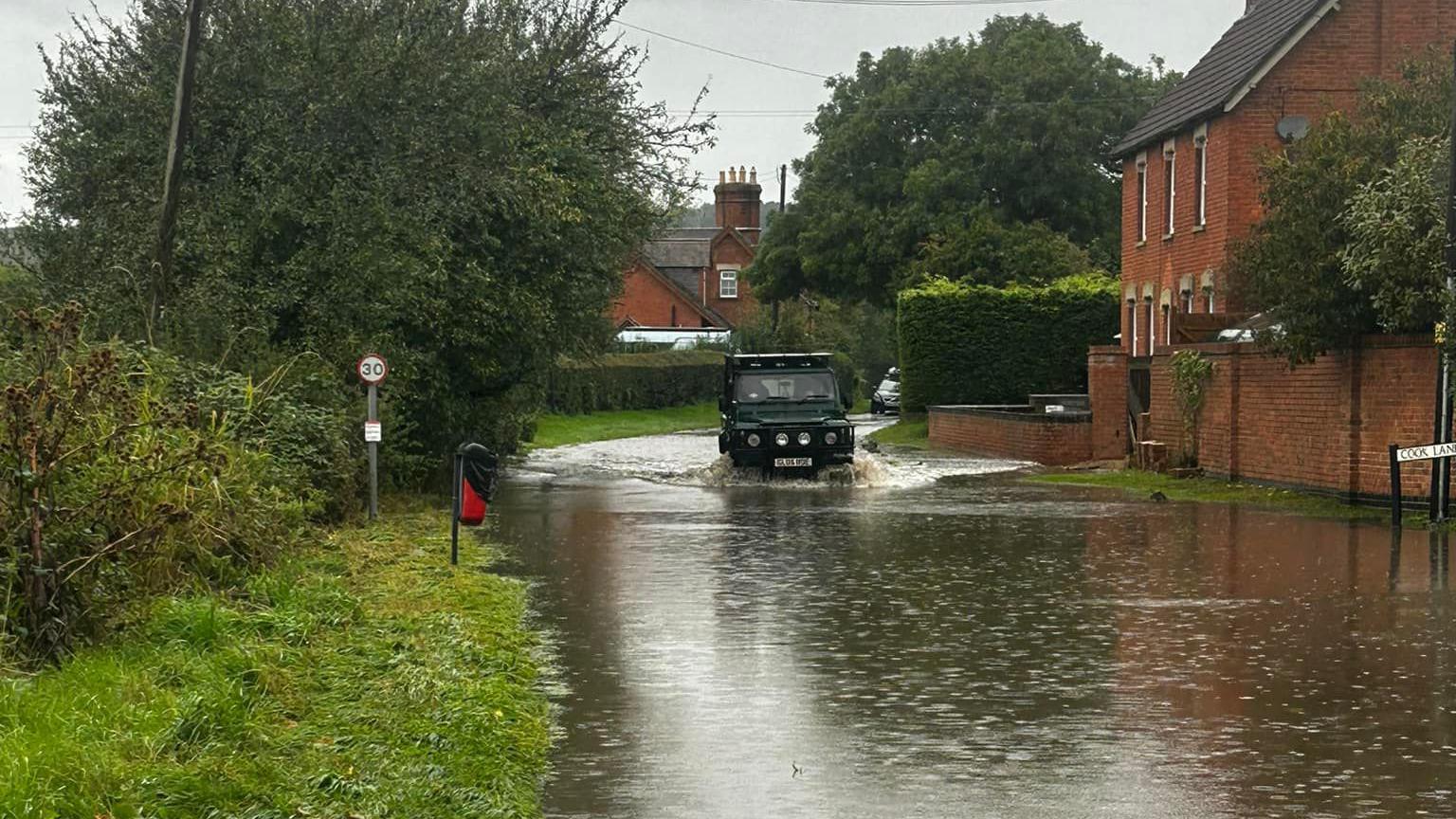 A 4x4 can be seen entering flood water. The water looks deep and is speckled with rainfall. Red brick houses can be seen to the rear and to the right. 