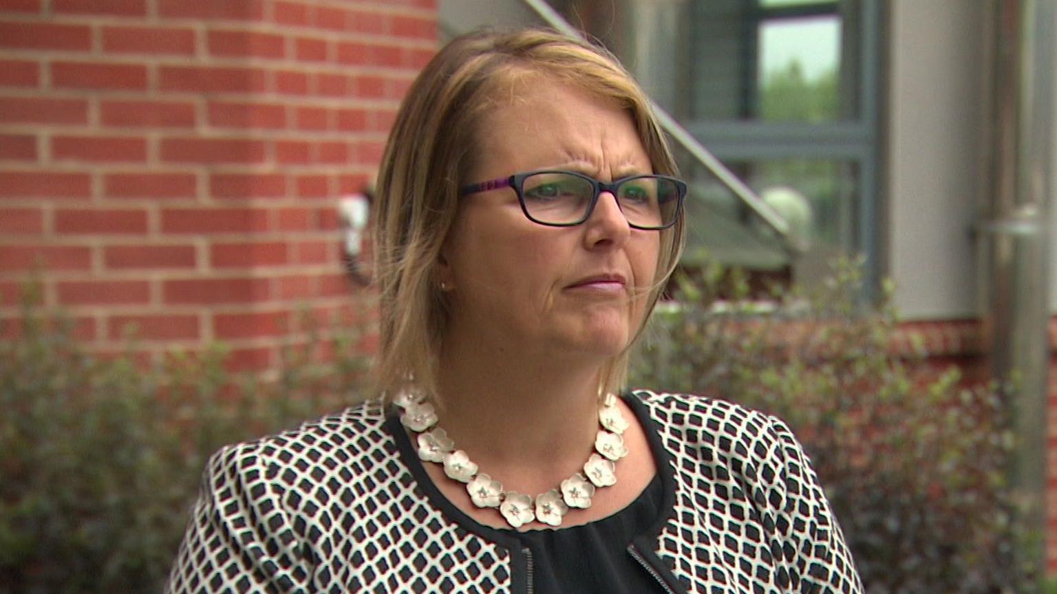A woman, Tina Graham, with medium length brown hair, in glasses wearing a black and white jacket and a flowery silver necklace