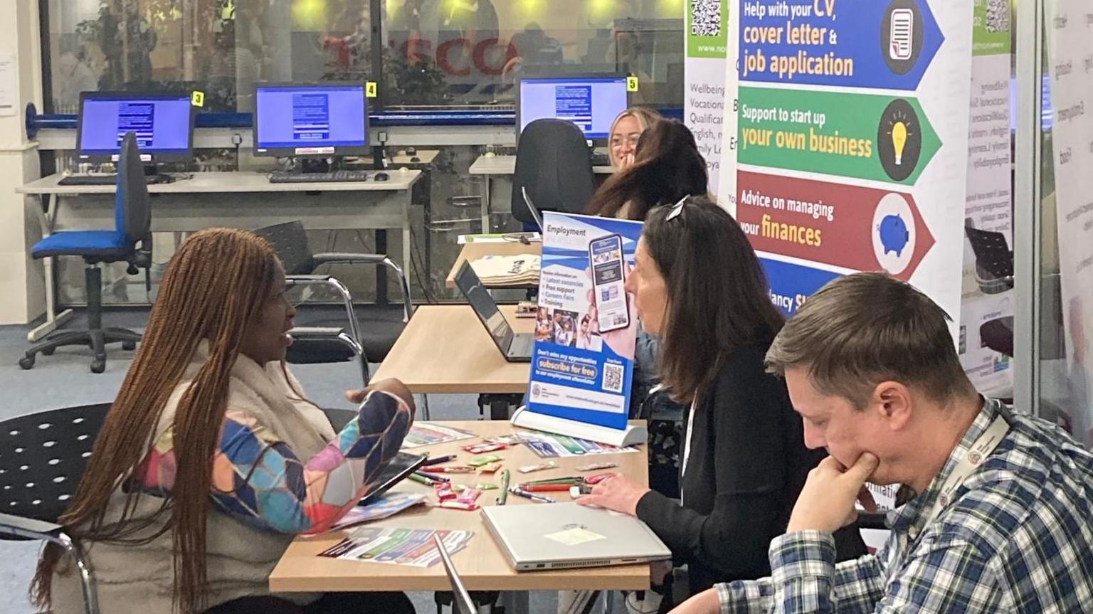 Two women sit at a desk covered in leaflets. There is a signpost to different services in the background.