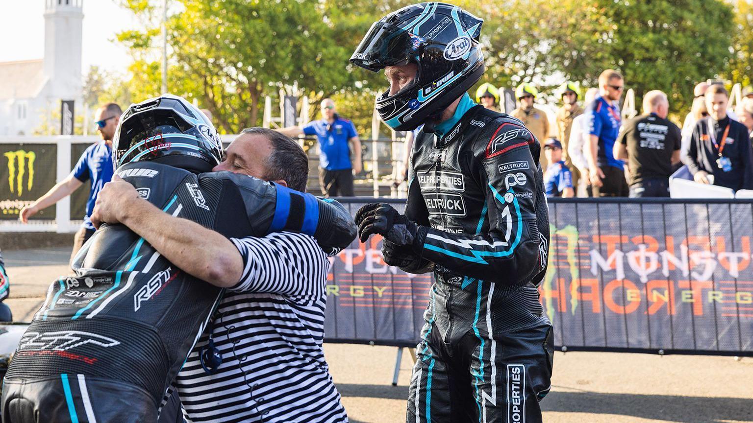 A man dressed in black motorcycle leathers and a helmet hugging a man in a black and white stripped T-shirt, who has  another man in the same colour leathers standing behind him 