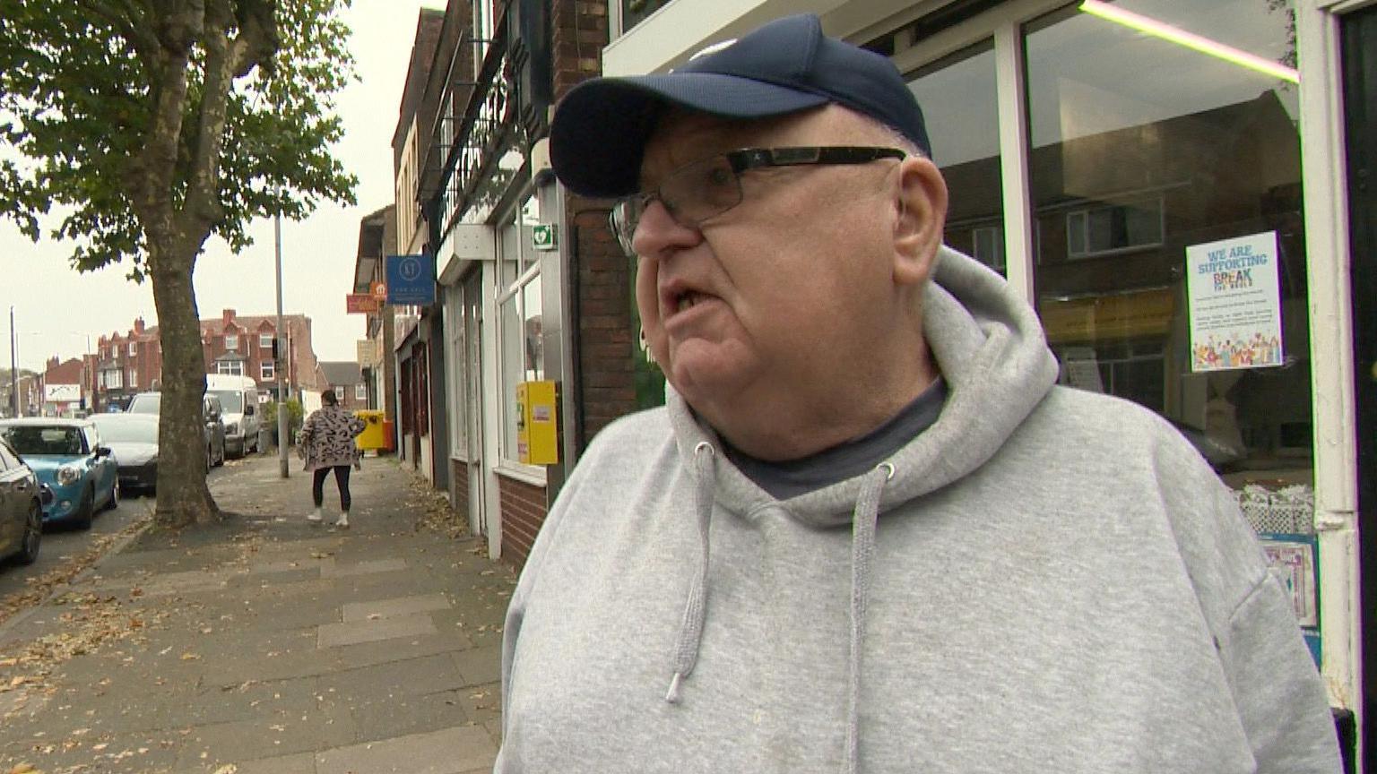Tony is wearing a grey hooded top, a blue baseball cap and black-framed glasses. He is standing on a Wirral high street, on the pavement in front of shops. There are shops and a number of parked cars behind him as well as a tree.