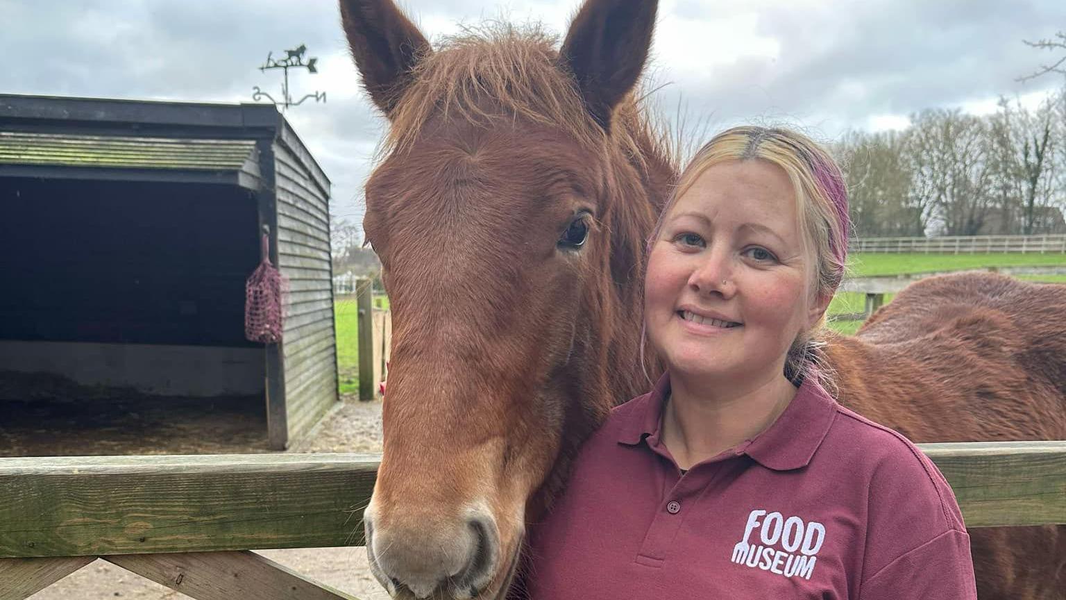 Melissa Abbott smiles at the camera while standing next to a Suffolk Punch chestnut coloured horse. Ms Abbott has blonde hair with some dyed pink parts that is tied back. She is wearing a burgundy coloured polo top with the words "Food Museum" written on the chest. Behind her is a field with an outbuilding for the horse. 