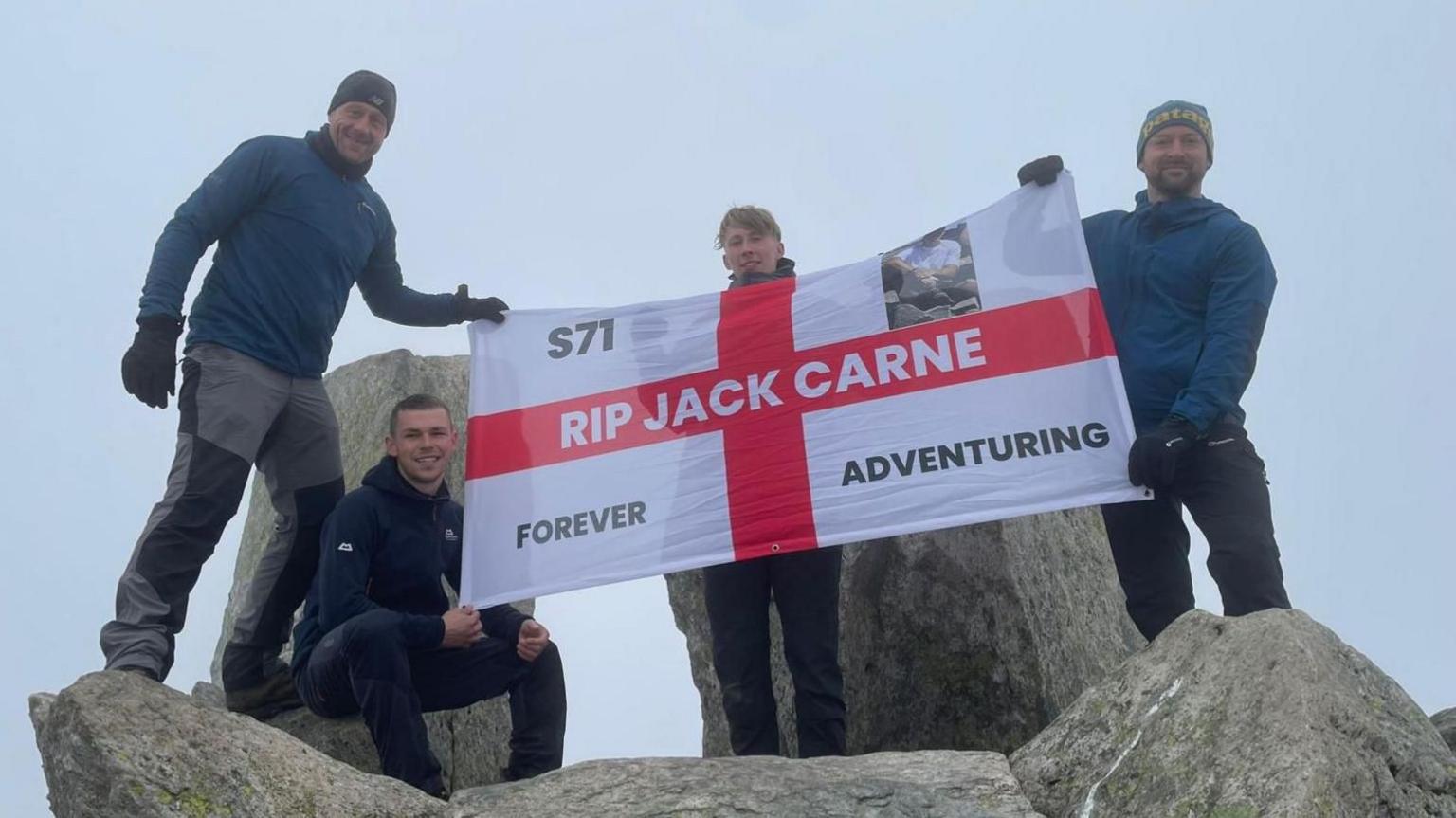 Pedwar dyn yn sefyll ar ben creigiau llwyd mawr yn dal baner sy'n nodi 'RIP Jack Carne', a 'forever adventuring'