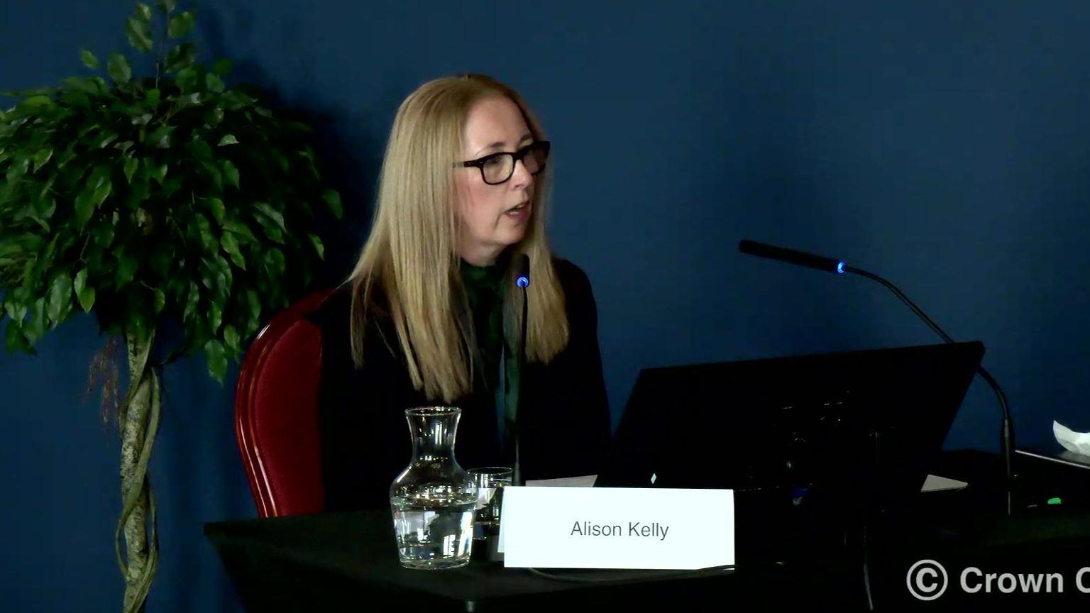 Alison Kelly, who has shoulder length reddish blonde hair and wears dark rimmed glasses and a green top under a black jacket, sits at a desk with a computer screen, microphone and a notice with her name on 