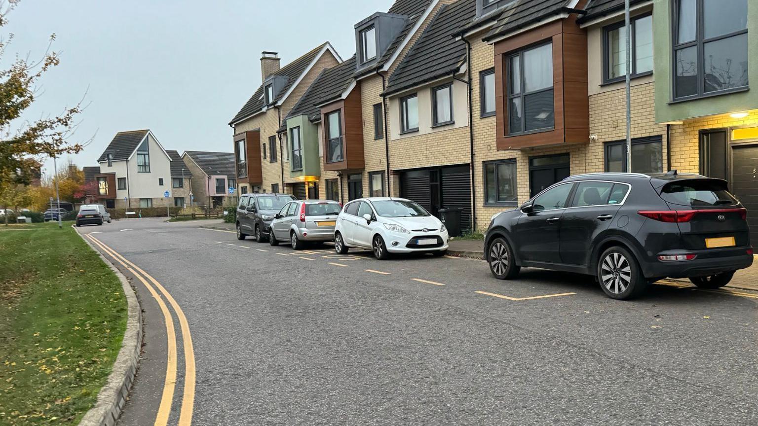 A bus stop with three cars and one van parked across it. The bus stop is painted on the road outside a row of terraced houses.