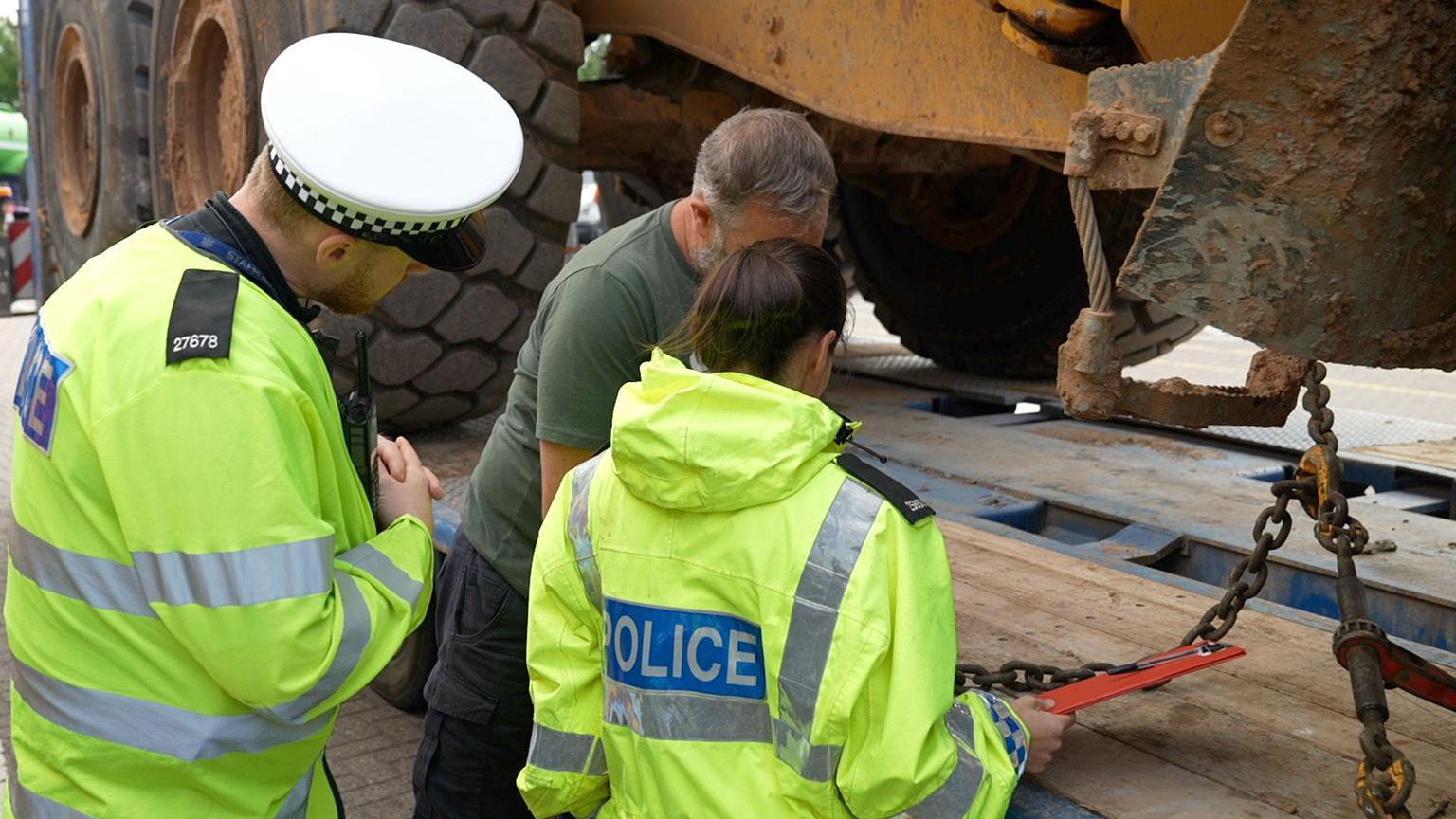 A man and a woman in high vis yellow jackets with a blue police badge on the back stand with a man in a green tshirt and navy cargo trousers in front of trailer carrying a vehicle with large tyres. 