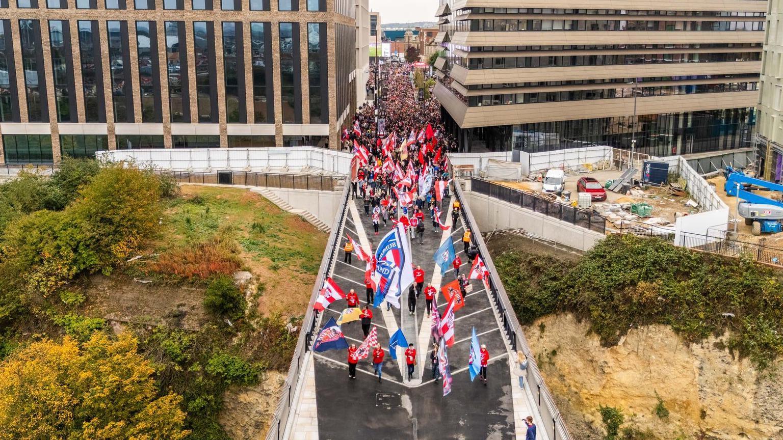 Overhead view of Sunderland fans on the new Keel Crossing. They are holding aloft large flags, and a mass of people can be seen behind them. To the right of the bridge building works are going on, with trees in autumnal colour to the left, and there are large buildings commercial buildings behind.