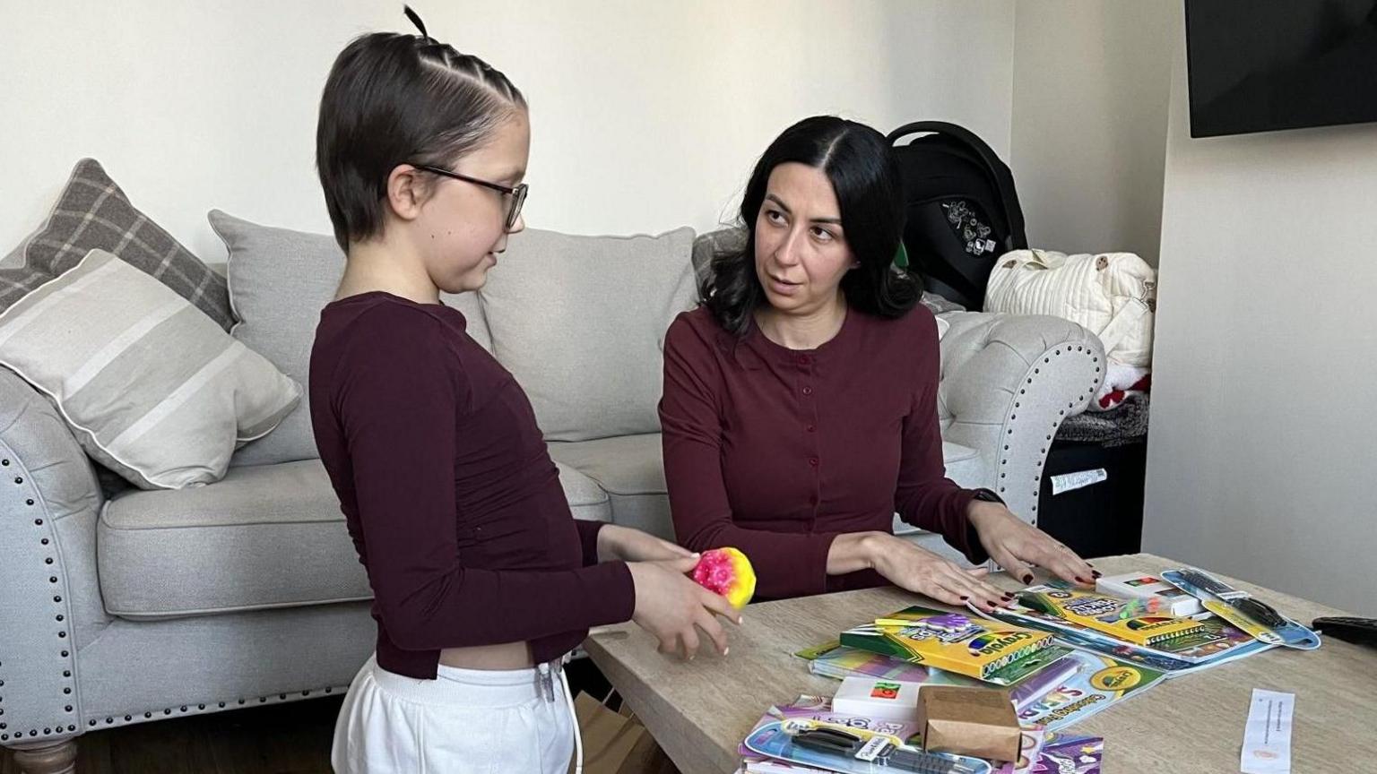 Mae merch 10 oed gyda gwallt tywyll byr, ac sy'n gwisgo top coch tywyll yn penlinio wrth fwrdd coffi pren sydd wedi'i orchuddio â phennau ffelt a llyfrau lliwio. Mae bagiau papur brown ar y llawr wrth ei hymyl. Mae menyw gyda gwallt tywyll hir a thop coch tywyll yn penlinio ar ben y bwrdd yn edrych ar y plentyn.