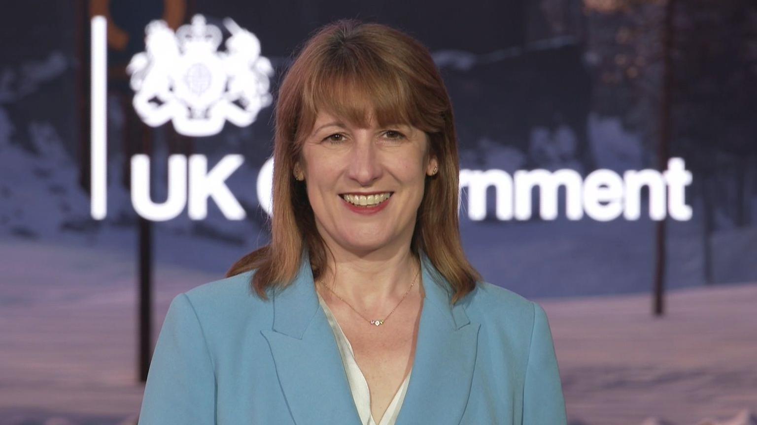 Rachel Reeves wearing a light blue suit with a white silk shirt and a gold necklace. She has shoulder length brown/red hair with a fringe. She is smiling in front of a screen that says UK Government.