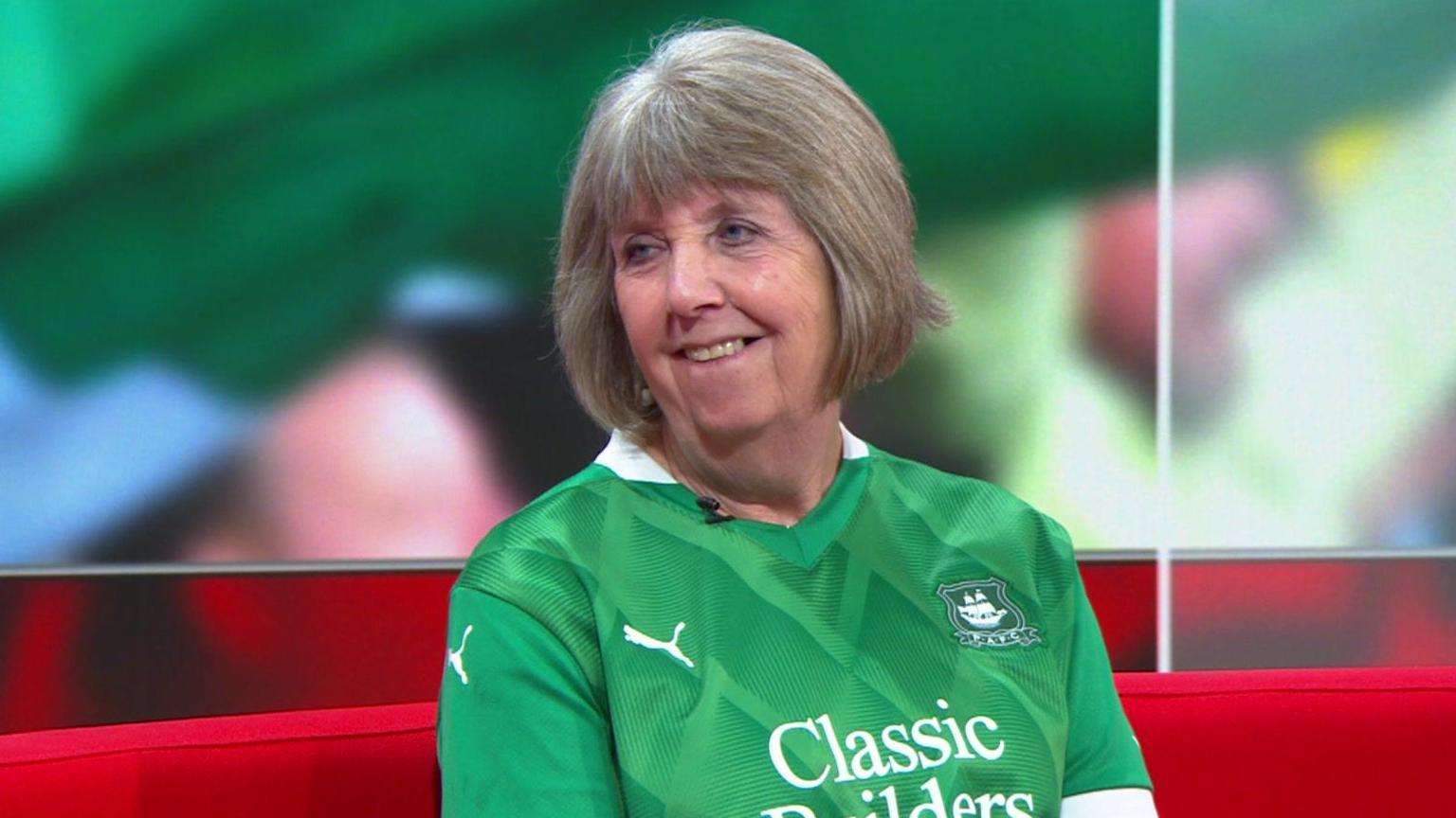 Julie Cardell sits on a red sofa in a TV studio. She is wearing a Plymouth Argyle home shirt, which is green in colour. She is looking to her right and is smiling with her teeth showing.