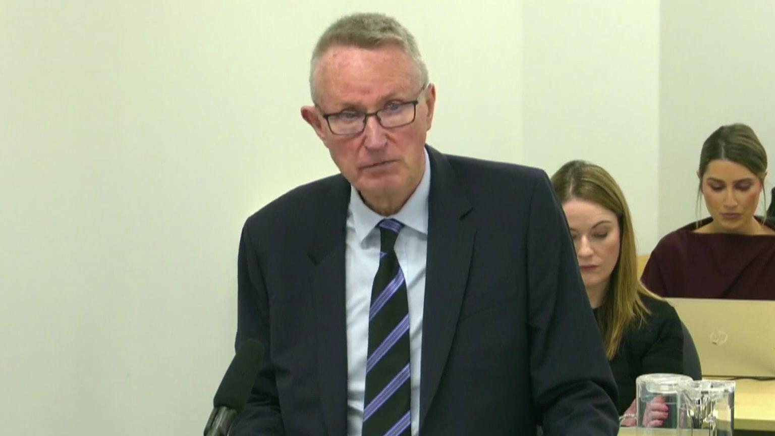 A man is standing in front of a mic. He's wearing a suit with a white shirt and a tripped black and purple tie. He wears glasses. Some people are sitting behind him. There are jugs of water on the tables.