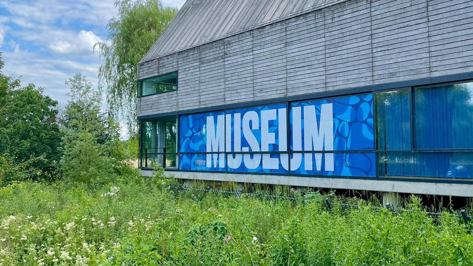 Outside the River and Rowing Museum in the sunshine with green plants all around a glass corner of the museum with the word "museum" on the side