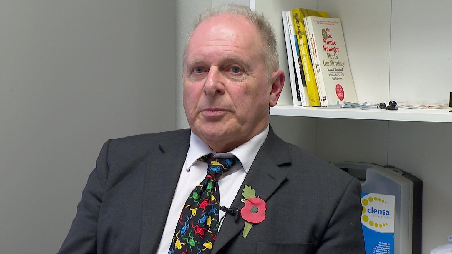 Defence solicitor Nigel Butt sits in front of a bookshelf. He has a black suit on with a white shirt and a multi-coloured tie. He has a poppy on his blazer's lapel.
