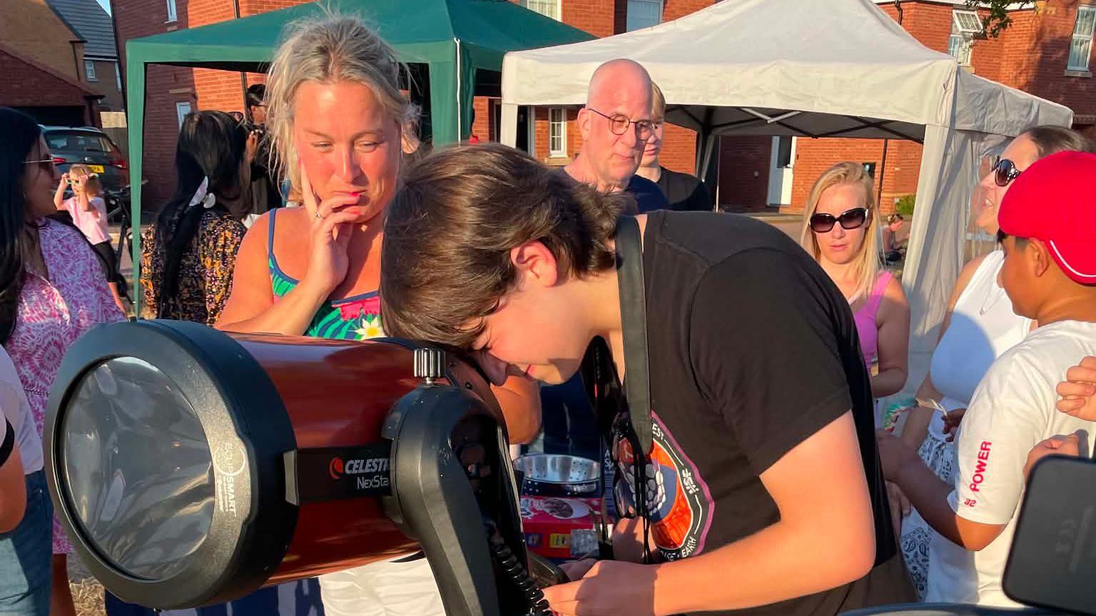 A young man with dark brown hair is looking through a telescope with a crod of people around him