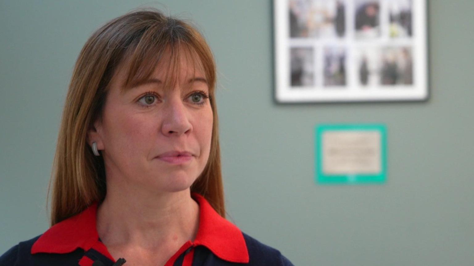 Sarah Suggitt has shoulder-length brown hair and is wearing a blue dress with a red collar. She is standing in the lounge of Elm House with a picture frame on the wall behind her