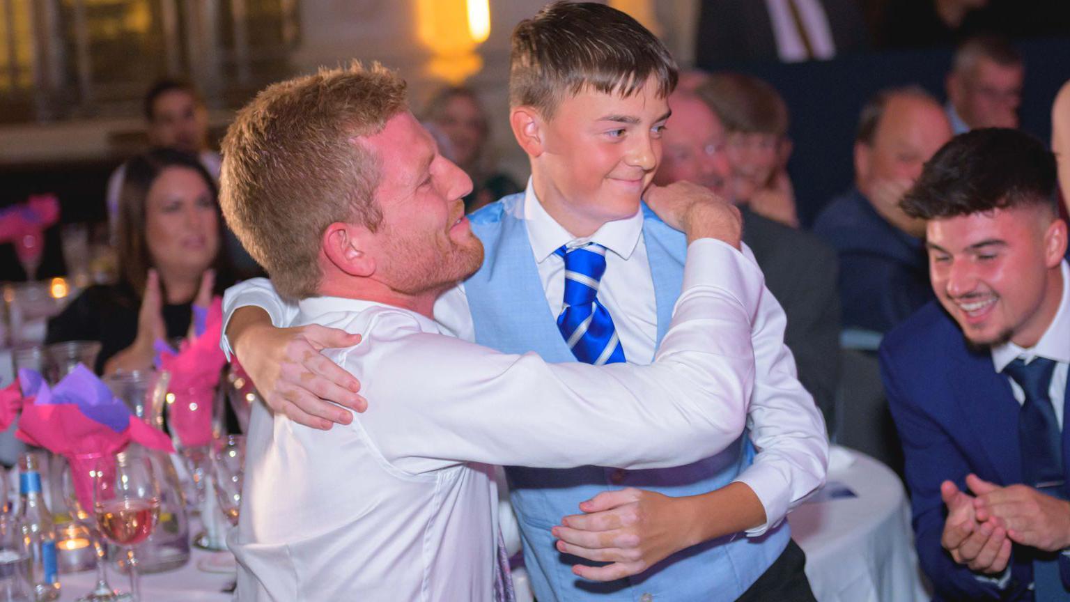 A boy wearing a blue waistcoat and tie and white shirt hugs his dad around a table whilst the audience clap for him in the background