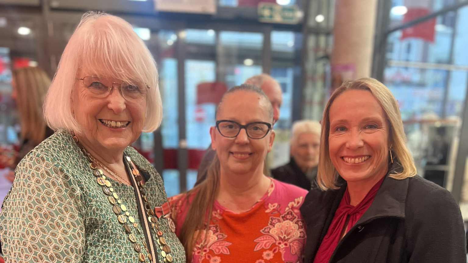 A woman with short white hair and wearing a green dress stands beside two women. One woman has brown hair that has been tied back and is wearing a red top with a floral design while the second woman has shoulder-length blonde hair and is wearing a black coat over her red top. The women are stood inside a building with a glass door behind them.