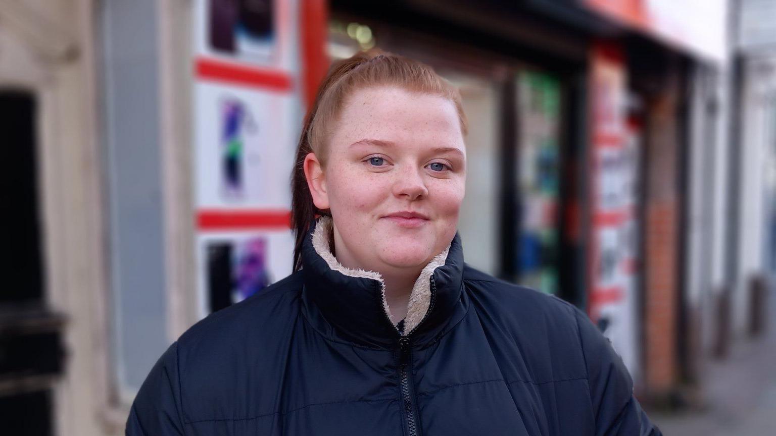 Woman with ginger hair and blue eyes smiling into the camera in a navy puffer coat with a white fluffy inside. 