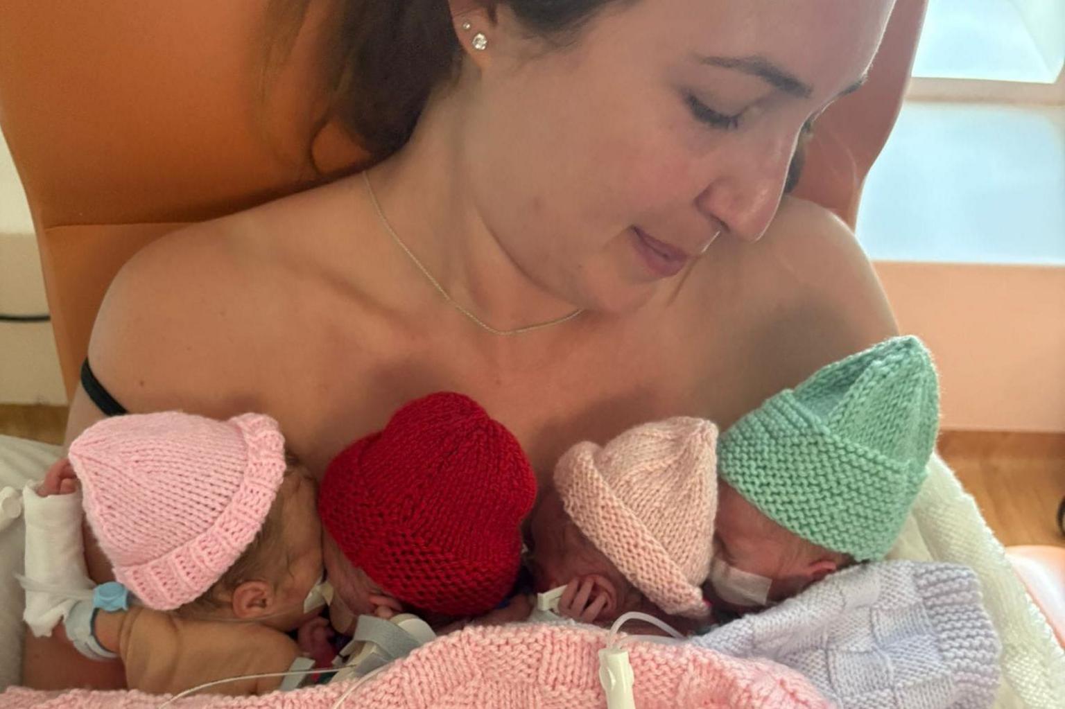 Jodie holds the four little babies to her chest during their time in care. They have assorted wires connected to them. They wear knitted hats and knitted blankets are around them.