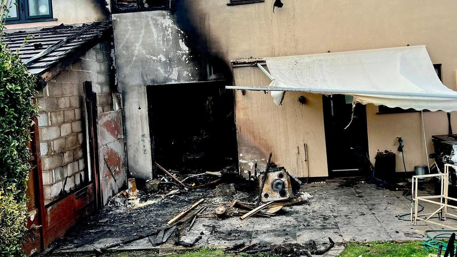 The outside patio of a house which has been ravaged by fire. The concrete is covered in black ash and burnt metal and wooden items. An awning above it is sagging and damaged.