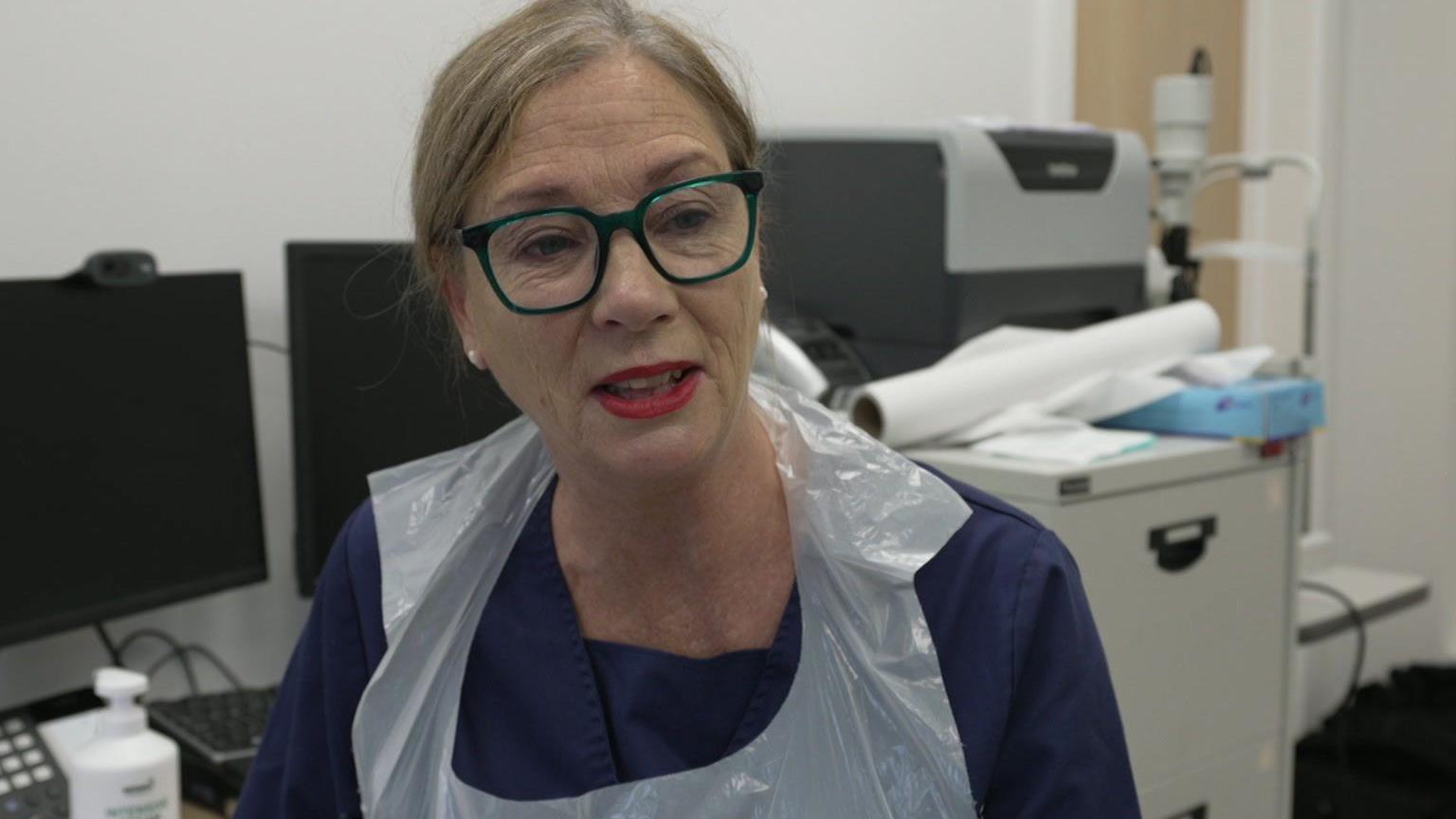 The picture shows a person wearing glasses and a plastic apron, seated in an office or medical environment. Behind them are two computer monitors and various office supplies. Some rolled-up papers rest on a filing cabinet in the background.