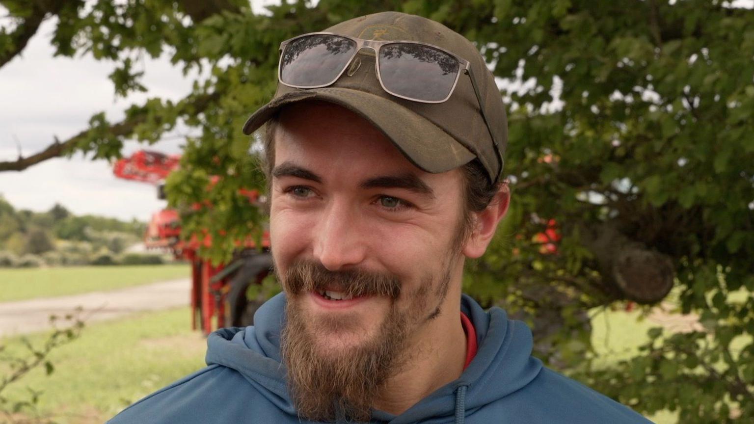 Morgan Hermon smiles as he looks away from the camera. He is standing outside near a field. A tree can be seen behind him as well as farming equipment. He wears a green cap with sunglasses resting on it as well as a blue hoodie. He has a long beard.