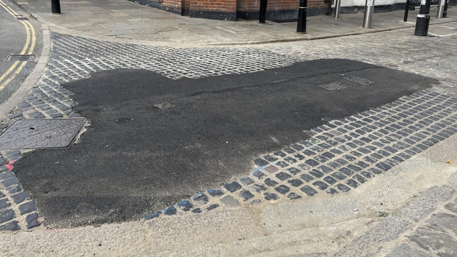 A section of a cobbled street which has had tarmac put on a section of it.