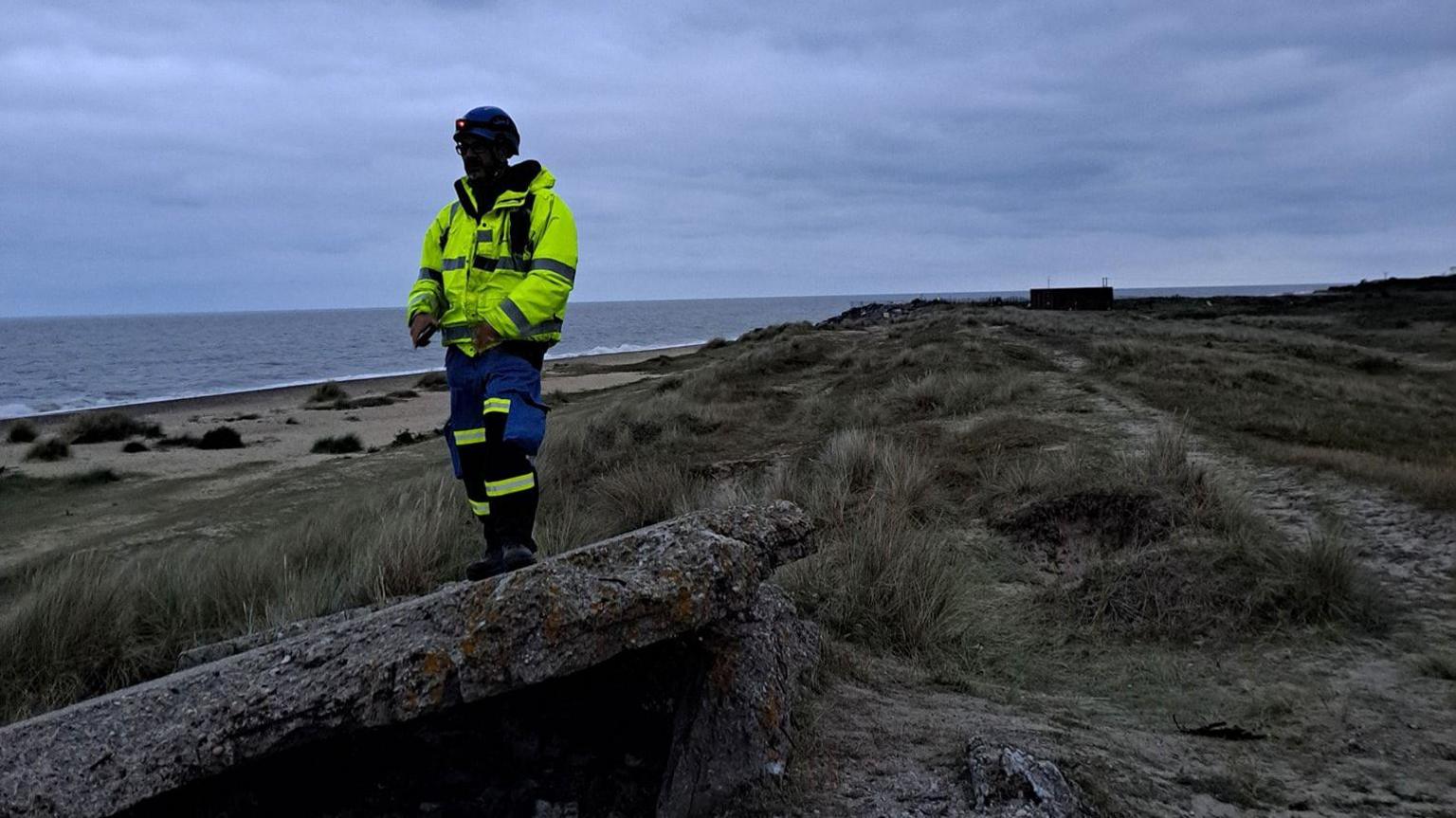 A person in a high vis yellow jacket is standing on a piece of rock next to the coast. The sea and beach can be seen in the background and it appears to be the early morning. The persons face is not visible. 