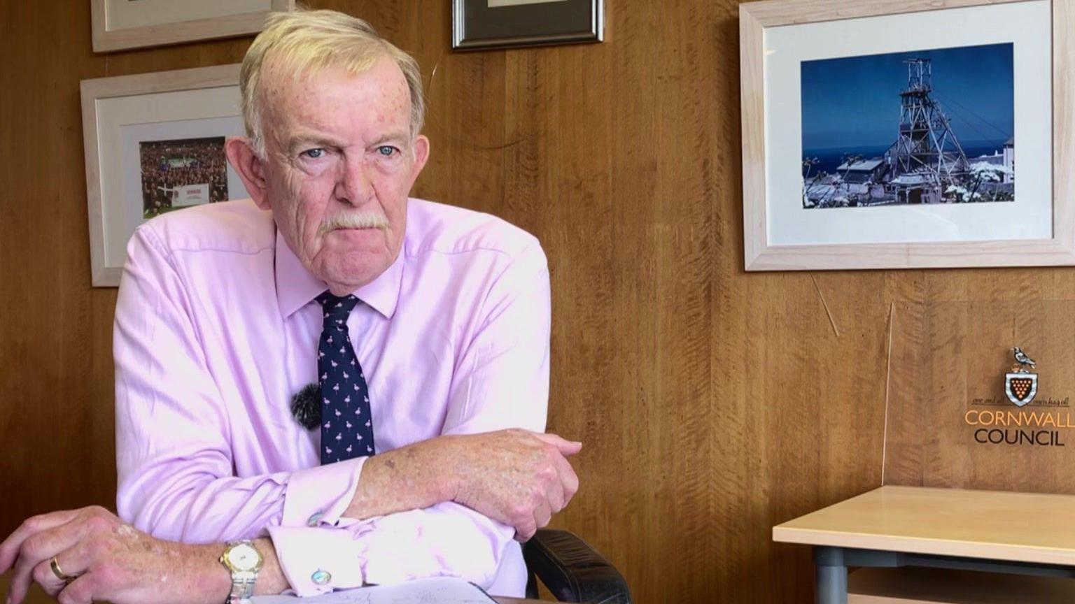 David Harris wearing a pink shirt and blue tie. He has grey hair and a moustache and sits at a desk in front of wood panelled wall with picture frames hanging on them