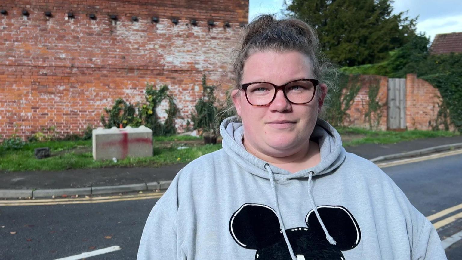 A woman with her hair in a messy bun, glasses and a grey hoodie with a Mickey Mouse outline on it stands on a pavement. On the other side of the road, a concrete block with a red cross spray painted on it has been placed on a grass verge. There is a red brick wall with ivy growing all over it.