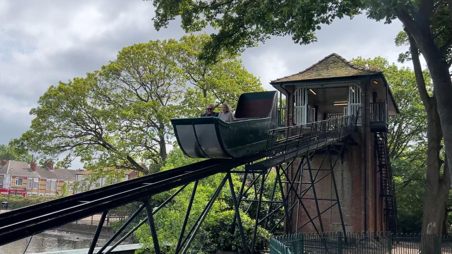 The red brick tower of the splash boat ride has a tiled roof and a metal flume extending down into the lake. The green boat is halfway down the slide and two figures are enjoying the ride