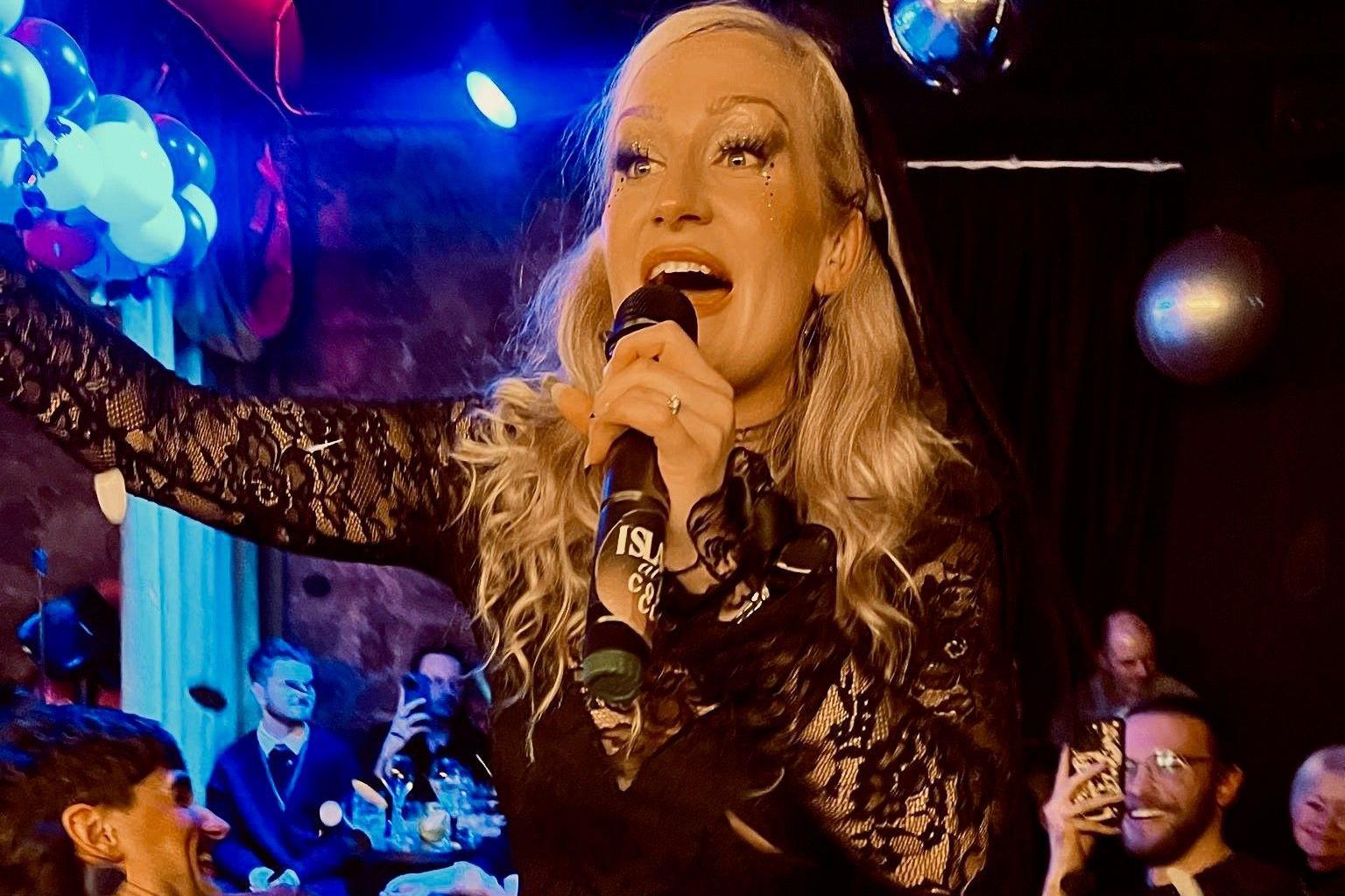 A blonde woman in a black top with lace sleeves sings into a microphone