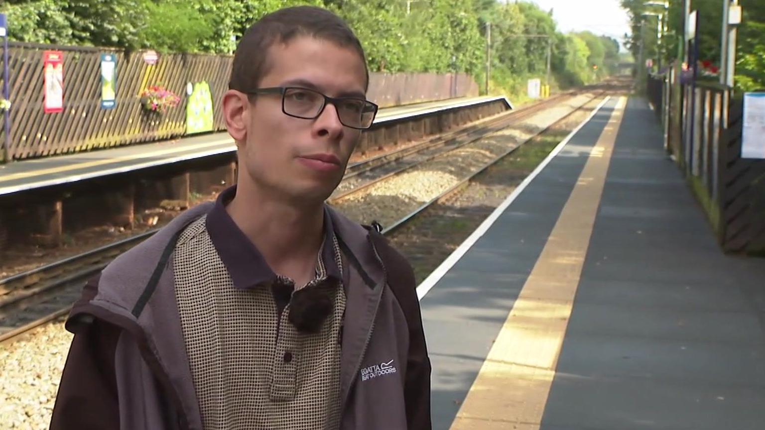 Nathaniel Yates is standing on the platform next to the railway line at Reddish North station. He is wearing glasses with a patterned shirt and a grey-and-black jacket. 
