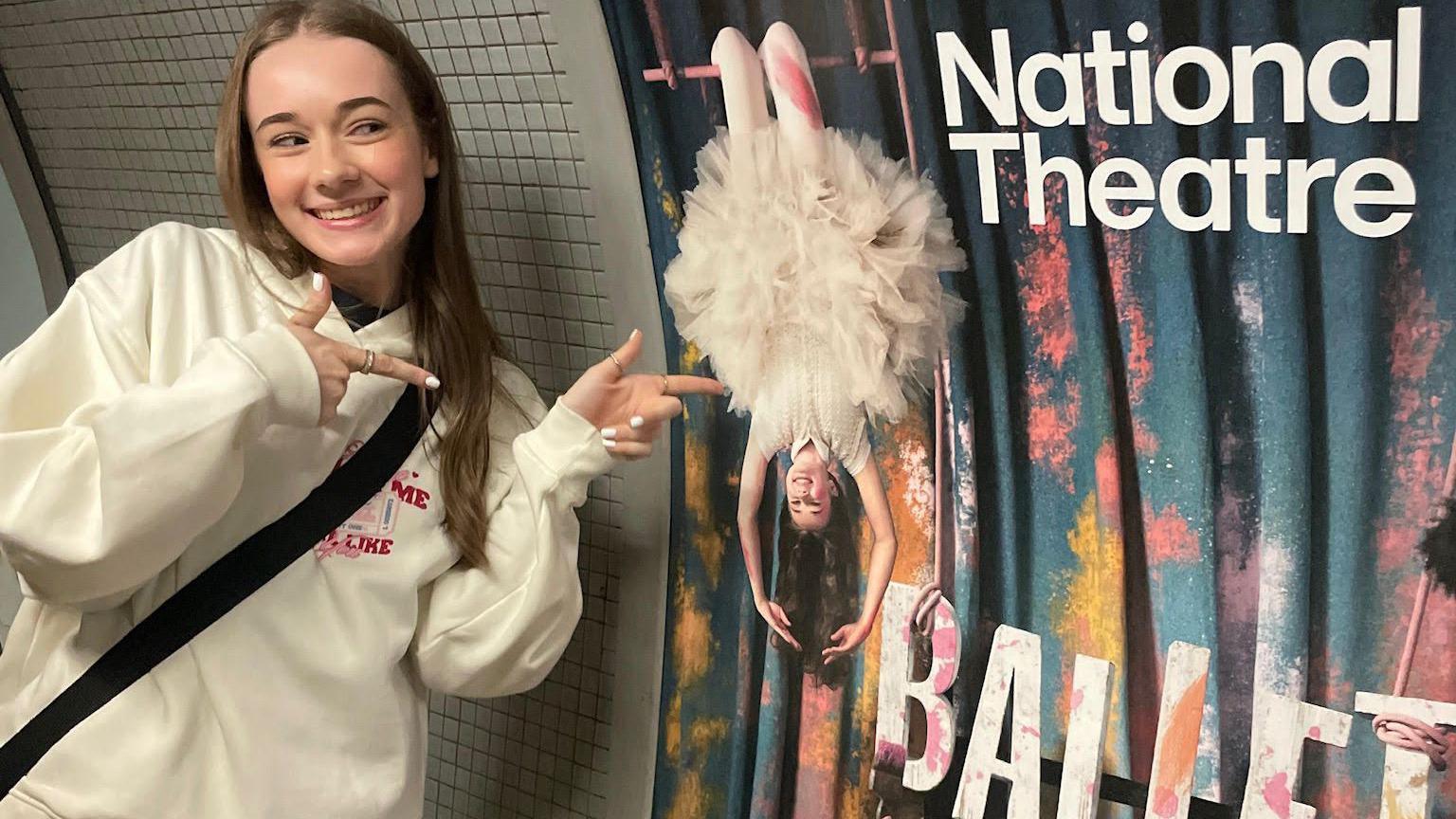 A young girl in a white hoodie is smiling and pointing to a large poster on a wall advertising a production.