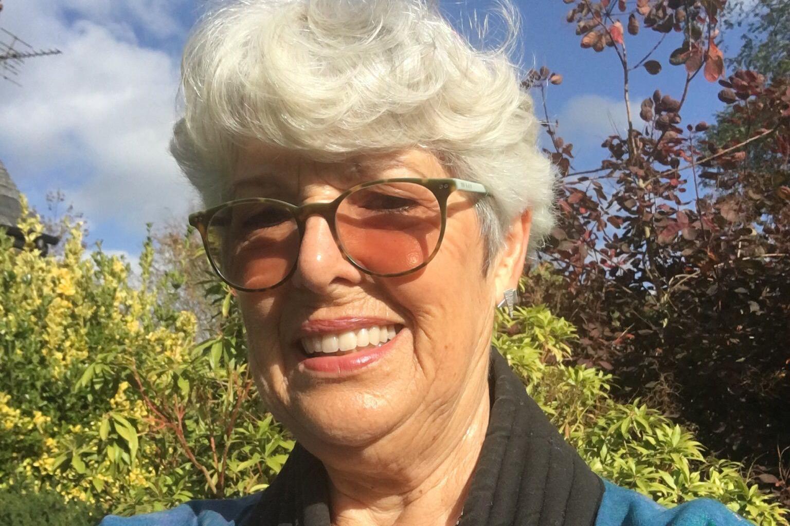 An elderly woman with short white hair and tinted glasses stands outside on a sunny day, smiling at the camera.