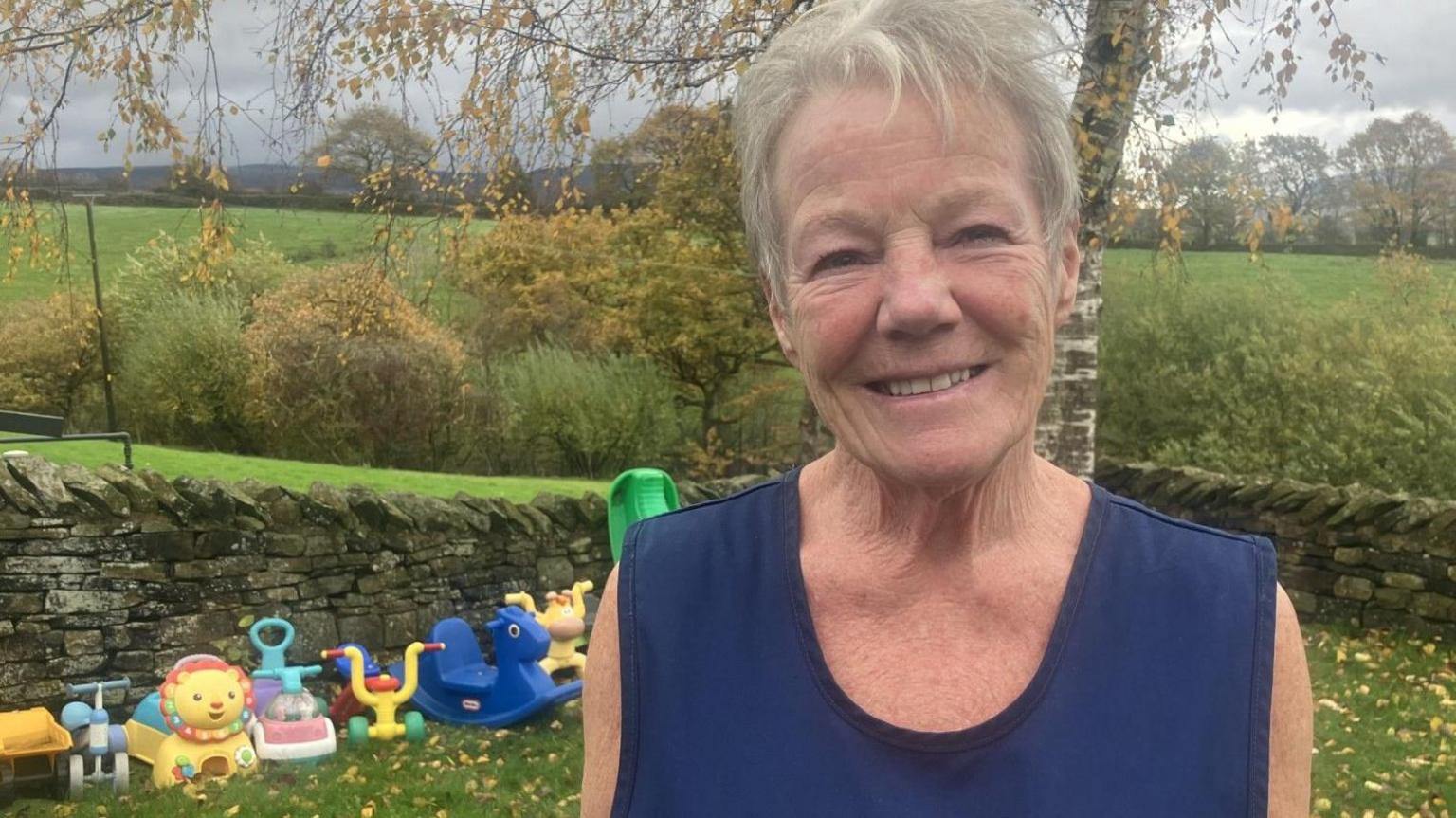 Maureen Savage, who owns a day nursery in the village of Hollingworth. With grey hair and wearing a navy blue top, she is pictured in the garden, with children's toys lined up against a stone wall in the background.