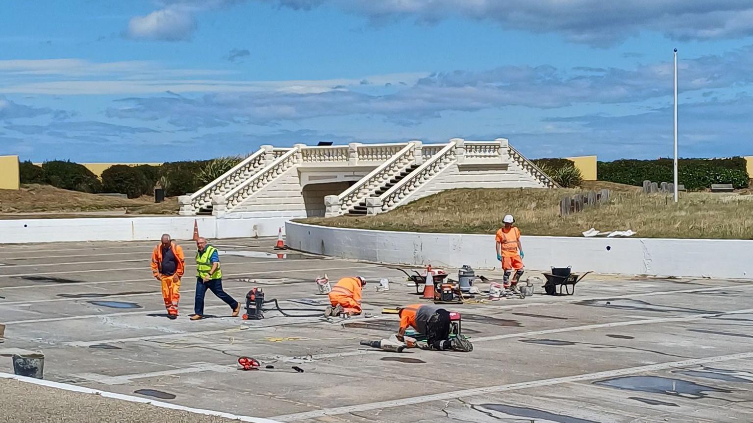Five men in high visibility orange clothing and white helmets are repairing the concrete floor of the lake, surrounded by building equipment and materials. There is a white ornamental footbridge at the back of the lake. The sky is blue. 