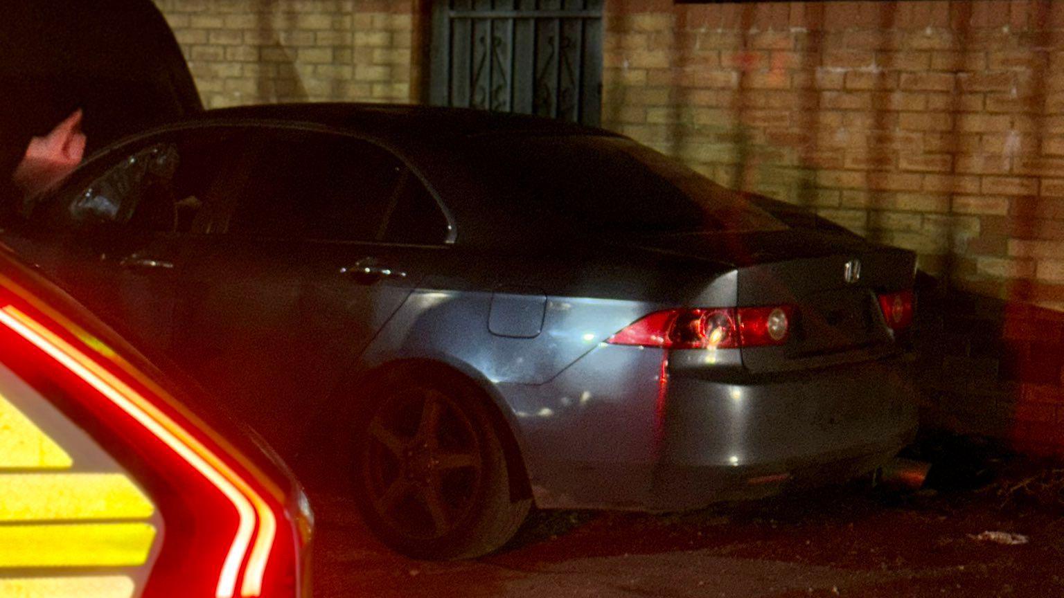 A dark car stopped by a brick wall, by a metal gate. A police car is to the left, with its lights on. It is dark and there are lots of shadows. 