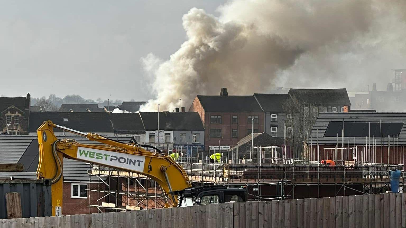 A large plume of smoke above a row of houses. There is a digger in the foreground and construction workers on a building site.