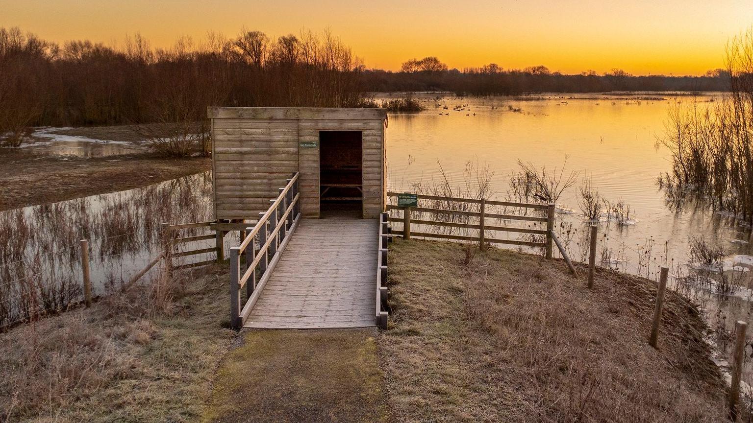 Small viewing platform and hide at the edge of grass and water course. It is dusk and the sky and lake is bathed in an orange glow.