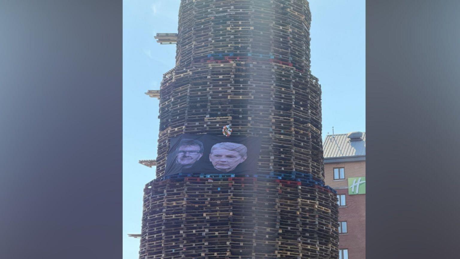 A towering bonfire built from wooden pallets stands on a city street on a sunny day. On the side of the bonfire there is a large black banner showing photos of the faces of Jeffrey Donaldson and his wife Eleanor. There is a red brick hotel in the background.
