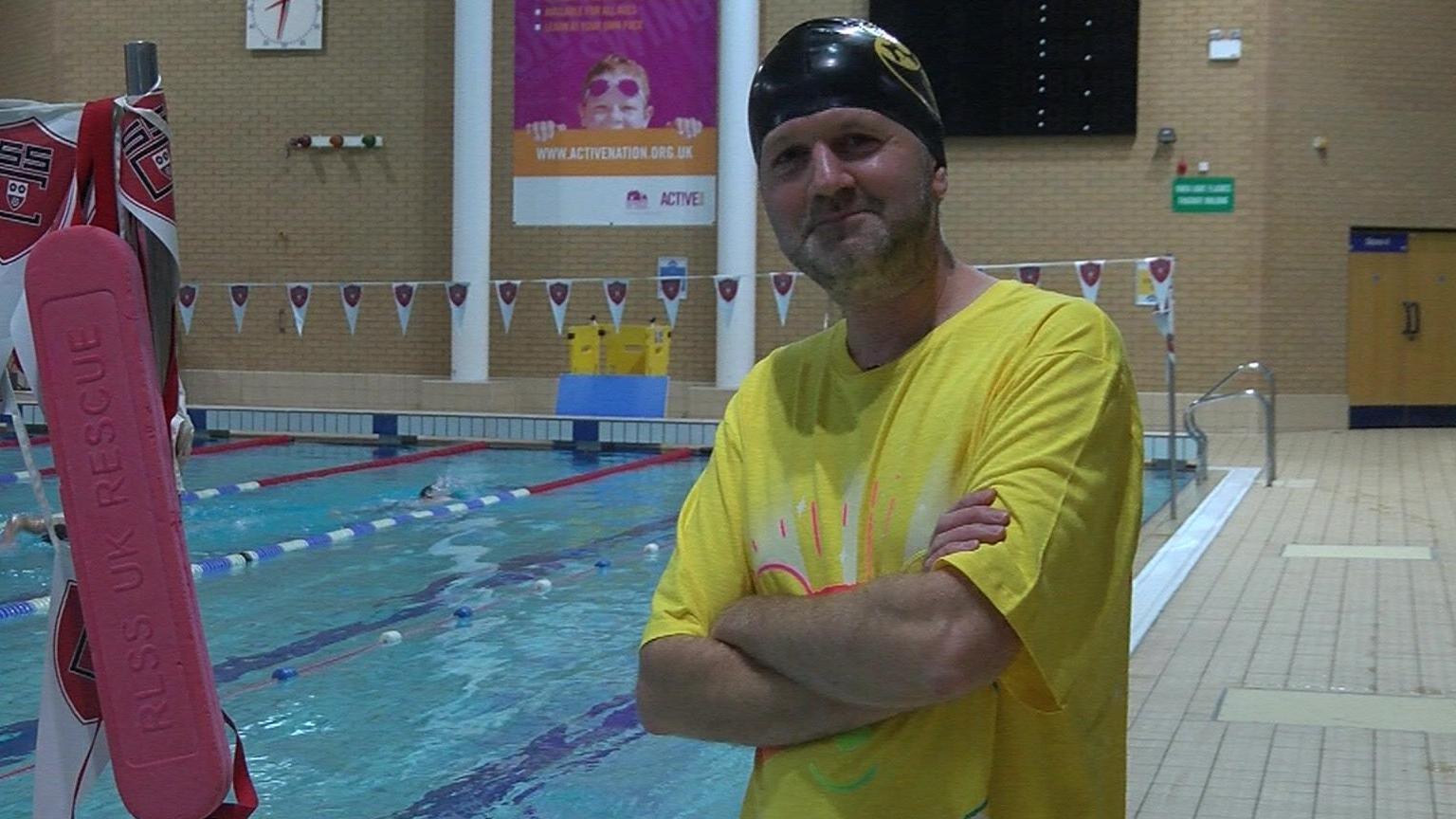 A man in a yellow t-shirt and a black swimming hat, standing in front of an indoor swimming pool, smiling with his arms folded