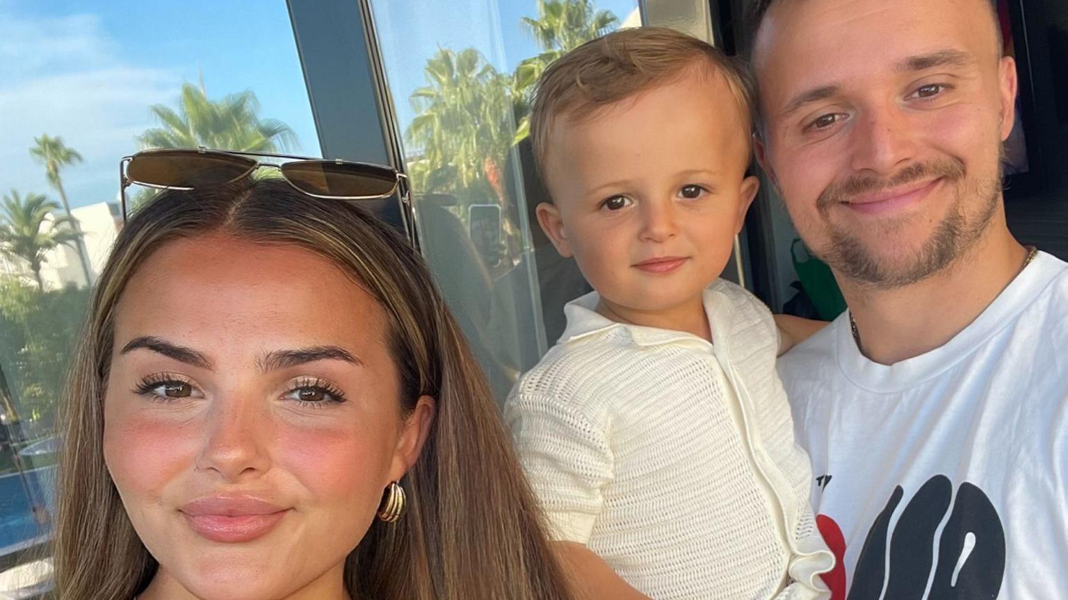 A family selfie photo of Chloe, Josh and their son Oakley, which appears to have been taken by mum Chloe. They are all smiling at the camera. Chloe has sunglasses on her head, and palm trees and a bright blue sky can be seen in the reflection of the window behind them.